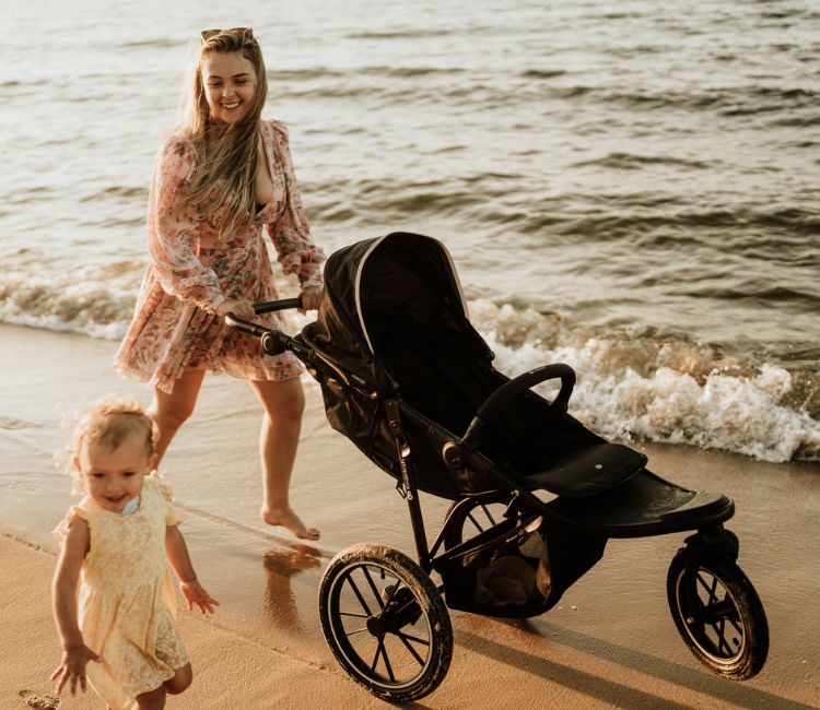 Mama prowadząca wózek HELSI marki Kinderkraft po plaży, kiedy jej dziecko samodzielnie idzie obok niej