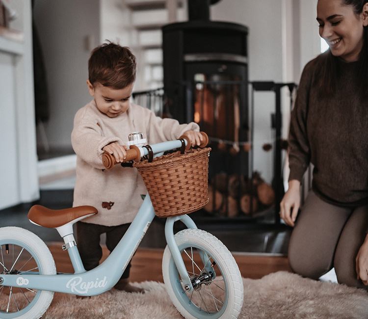 Dwuletni chłopiec bawi się rowerkiem biegowym w salonie, w towarzystwie matki, która jest w pobliżu.