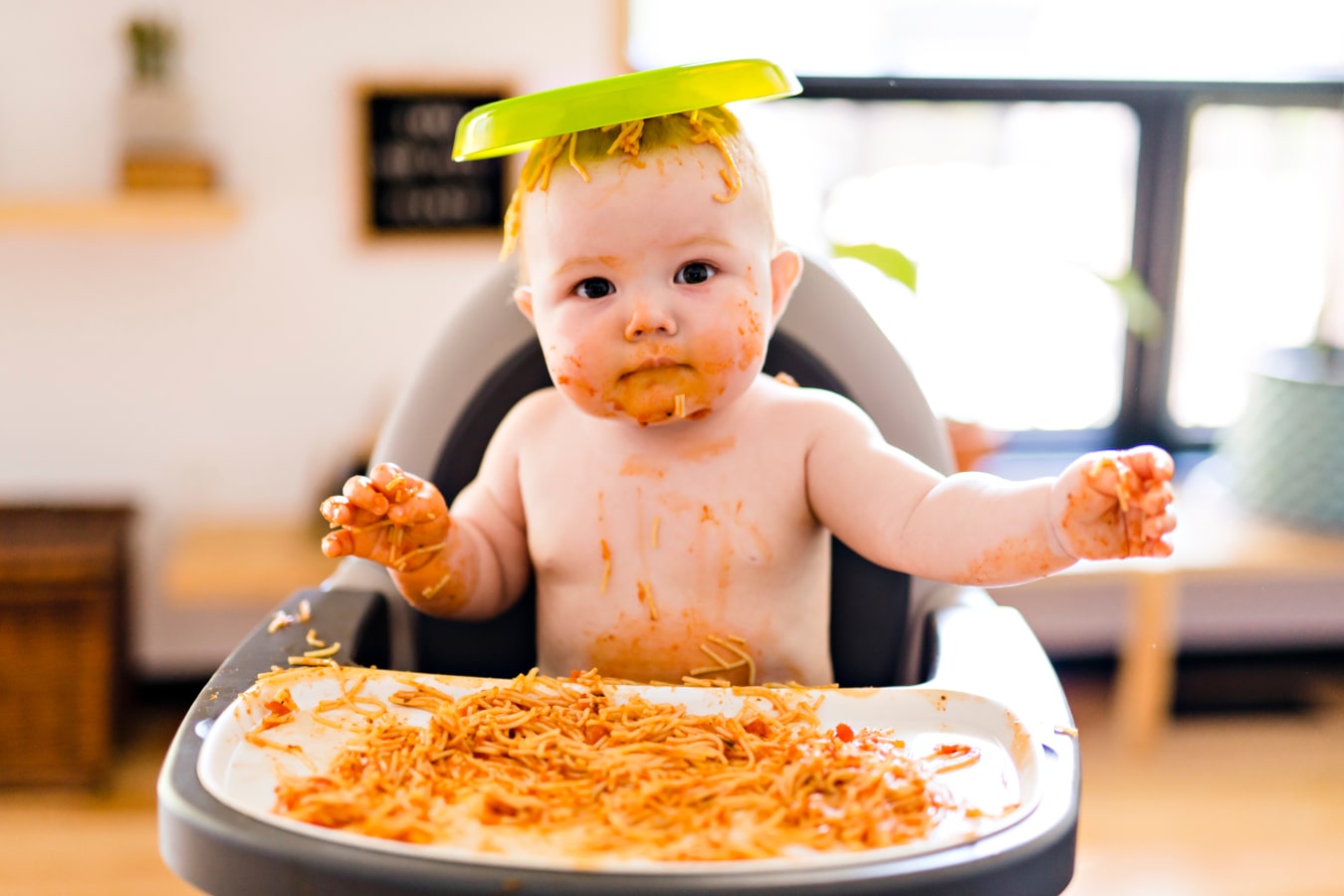 Brudne od spaghetti dziecko siedzi na krzesełku do karmienia, ma talerz  jedzeniem na głowie