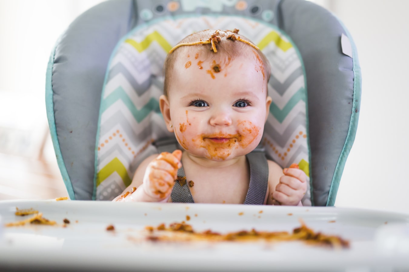 Brudne od spaghetti dziecko siedzi zapięte w krzesełku do karmienia. Jest szczęśliwe i się uśmiecha, na nim i na tacce jest pełno jedzenia.