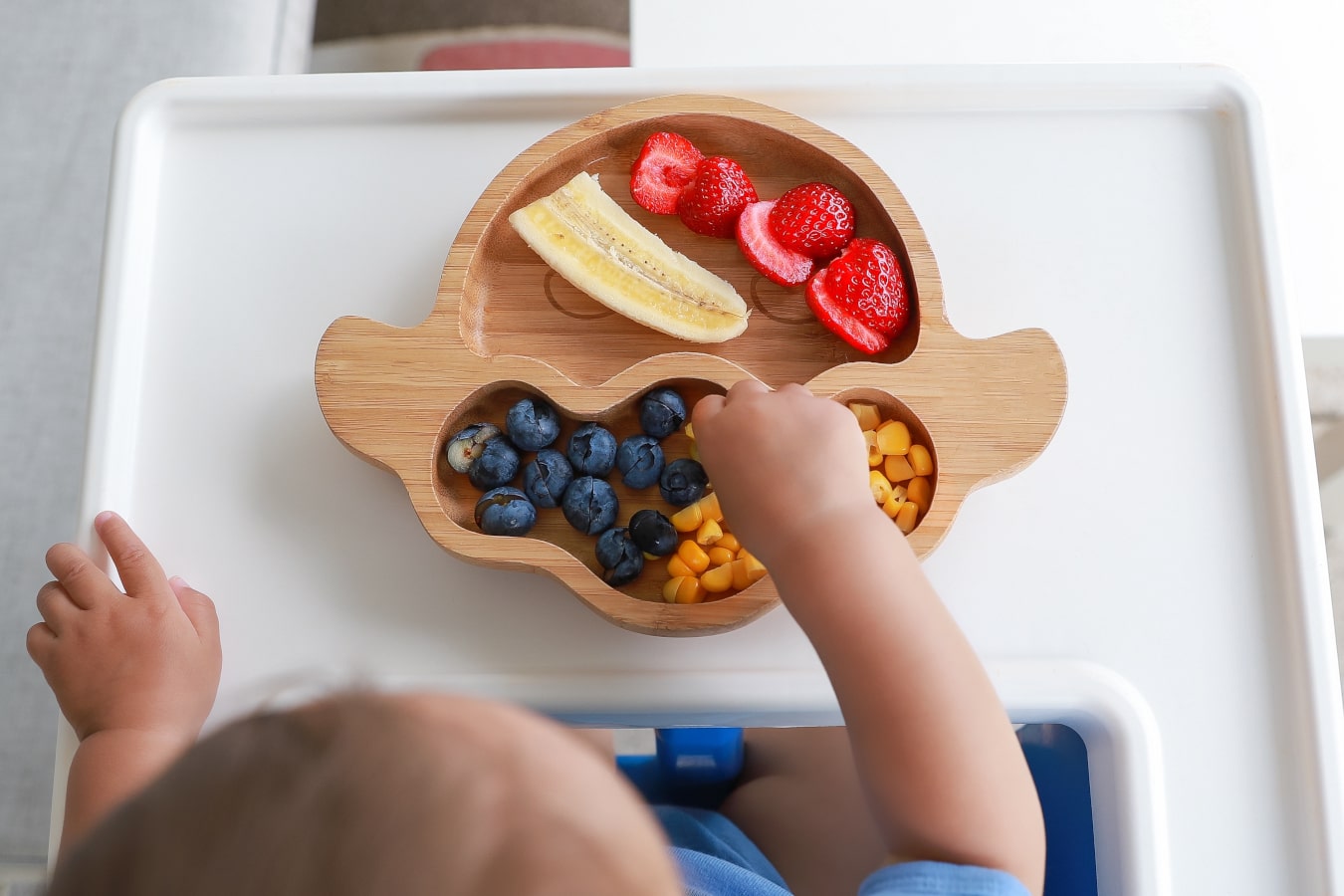 Na drewnianym talerzu z przegródkami widać kolorowe owoce i warzywa: banana, borówki, kukurydzę i truskawki. Dziecko sięga po jedzenie