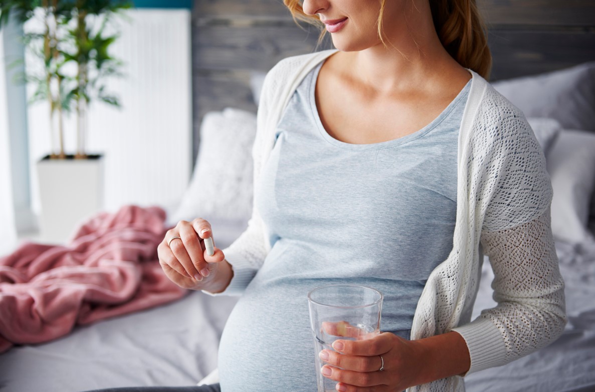 Kobieta w ciąży trzyma tabletkę i szklankę wody, ma widoczny brzuszek ciążowy