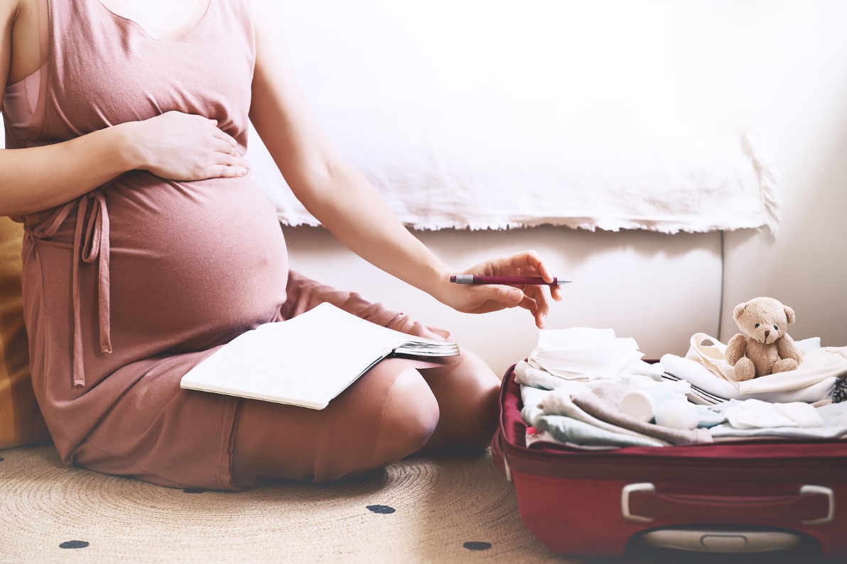 Kobieta w ciąży trzyma się za brzuch i pakuje walizkę do szpitala, na kolanach ma kalendarz