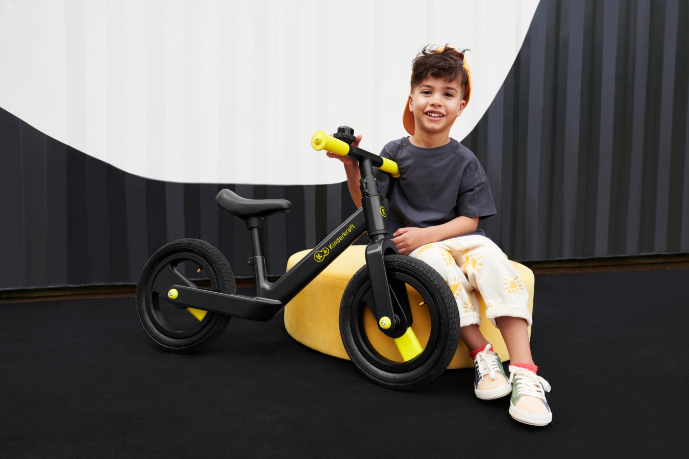 Wyluzowane dziecko siedzi na pufie w abstrakcyjnej przestrzeni i trzyma dwukołowy rowerek biegowy w kolorze czarnym 
