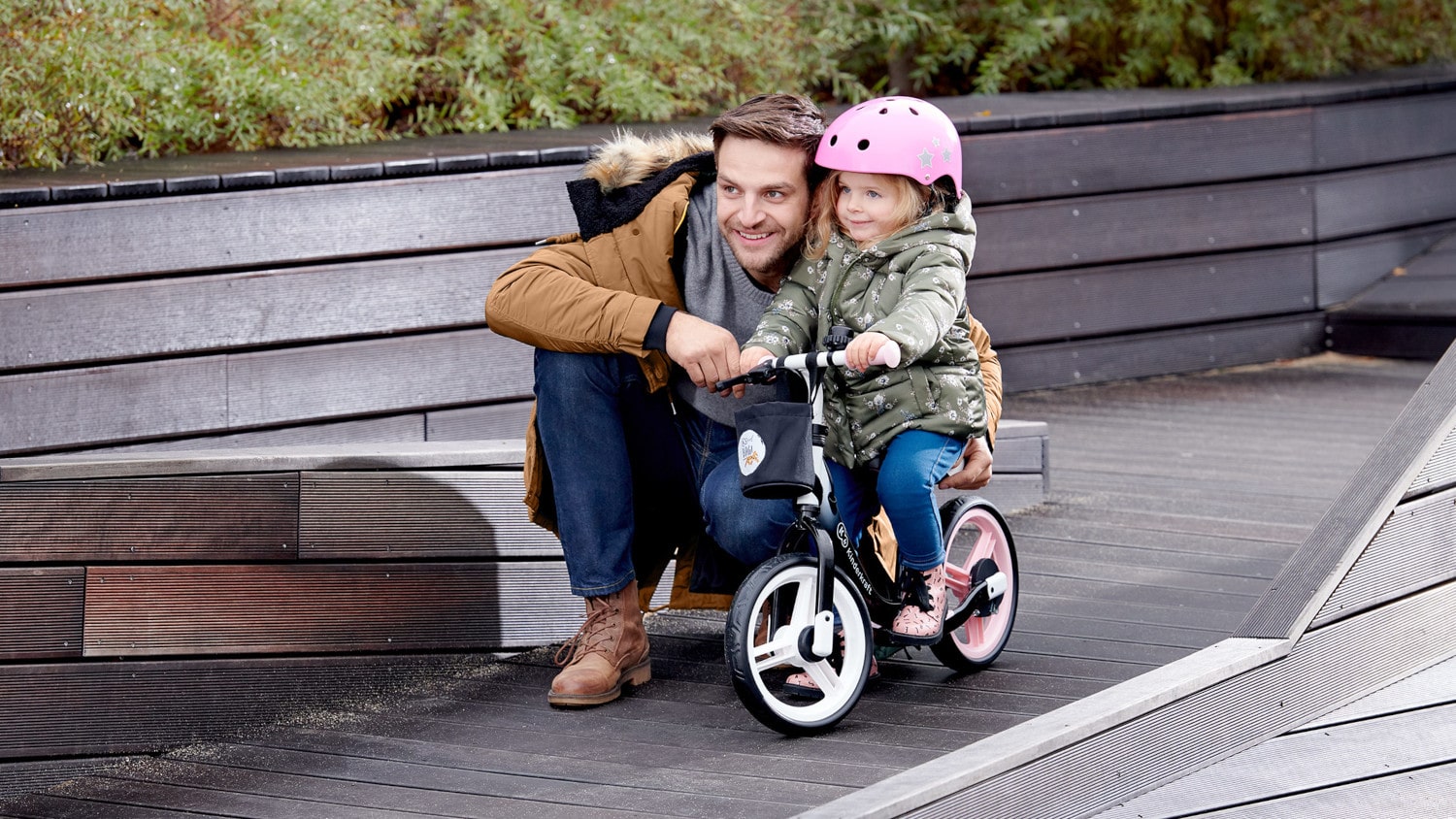 Ojciec na dworze jesienną porą kuca przy dziewczynce siedzącej w kasku na dwukołowym rowerku biegowym w kolorze czarno-różowym 
