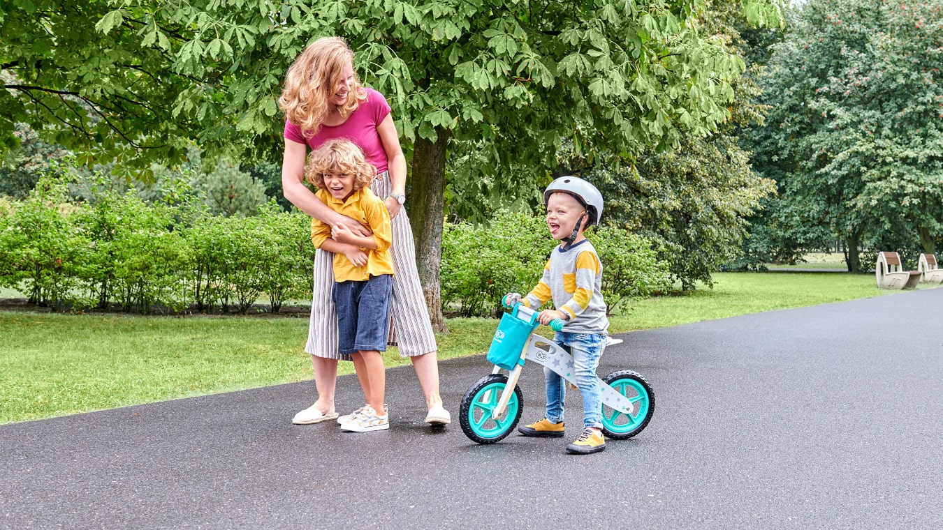 Mama spaceruje po parku z dzieckiem na dwukołowym rowerku biegowym w kolorze turkusowym i z drugim starszym dzieckiem 