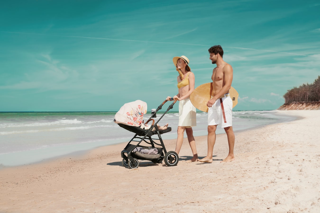 Mama i tata spacerują po plaży z dzieckiem leżącym w wózku spacerowym w kolorze czarnym z beżową budką 