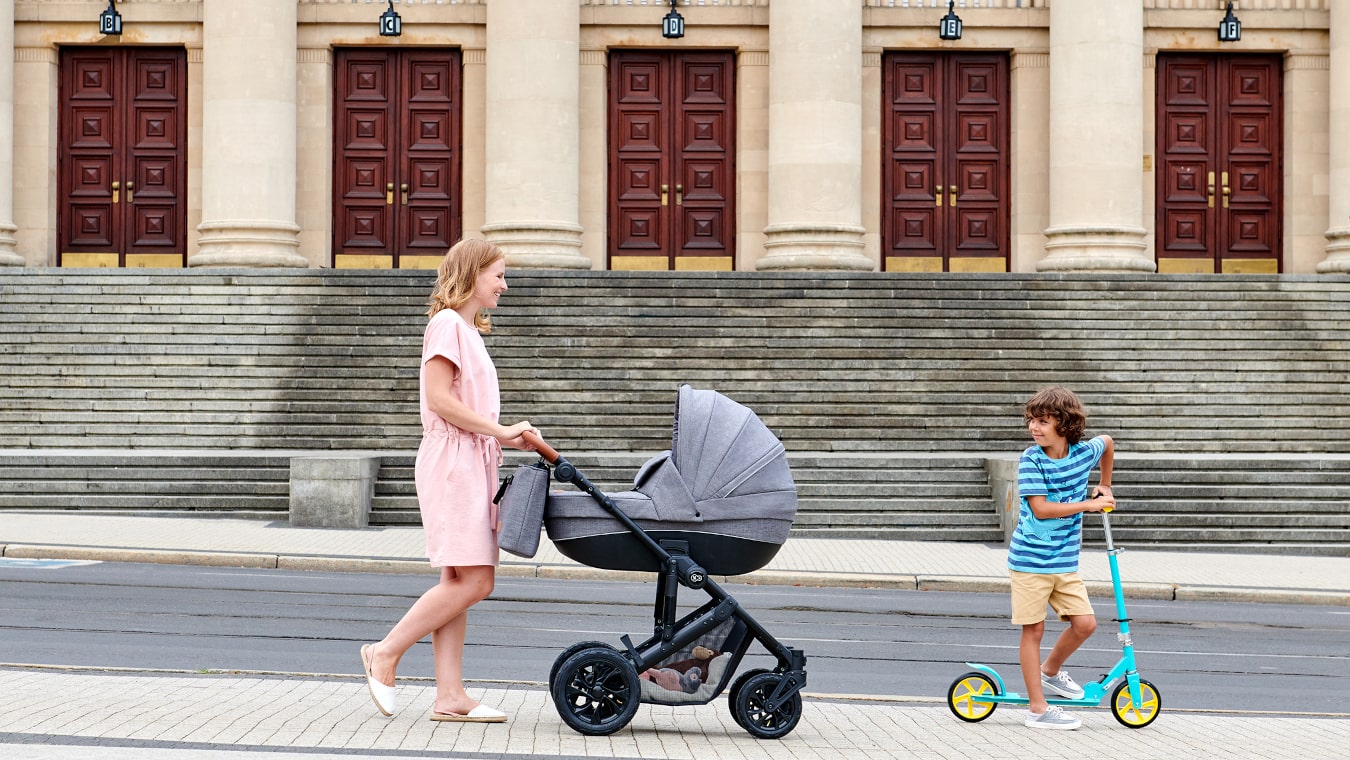 Mama spaceruje po mieście z głębokim wózkiem w kolorze szarym i z dzieckiem na dwukołowej hulajnodze w kolorze turkusowym 