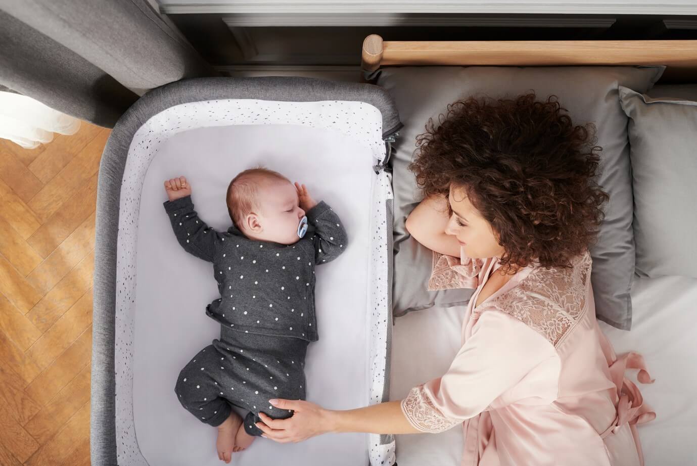 Ciemnowłosa kobieta leży na łóżku, obok niej śpi w dostawce dziecko. Kobieta uśmiecha się, dziecko w body z długim rękawem śpi spokojnie