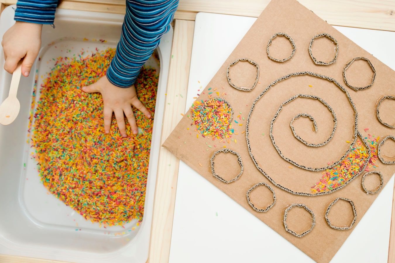 Rączki dziecka poznają nową fakturę - kolorowe cukierki i posypka, obok znajduje się kartonowy labirynt DIY