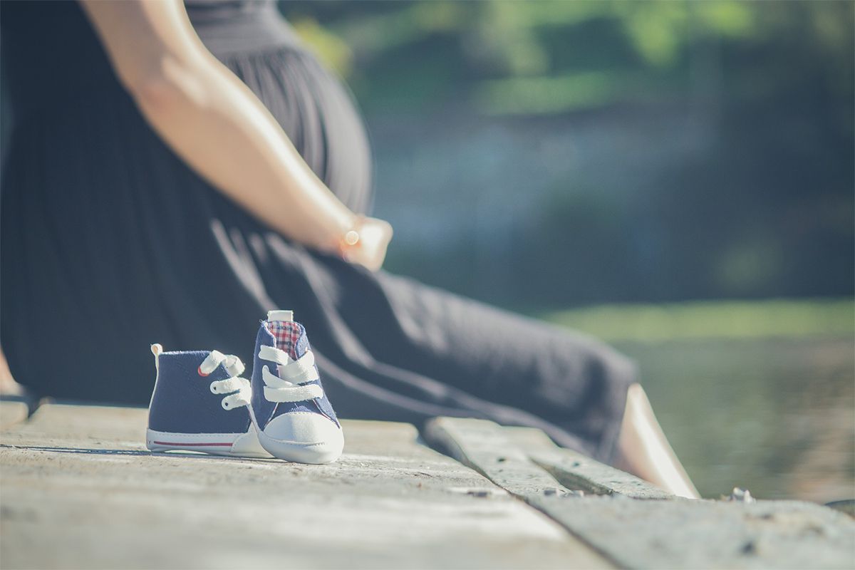 Dziecięce buty na ławce, kobieta w ciąży w tle, siedzi i trzyma się za ciążowy brzuszek
