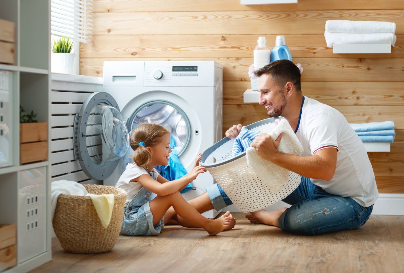 Roześmiany tata pokazuje córce kosz z praniem, razem doskonale się bawią przy pralce