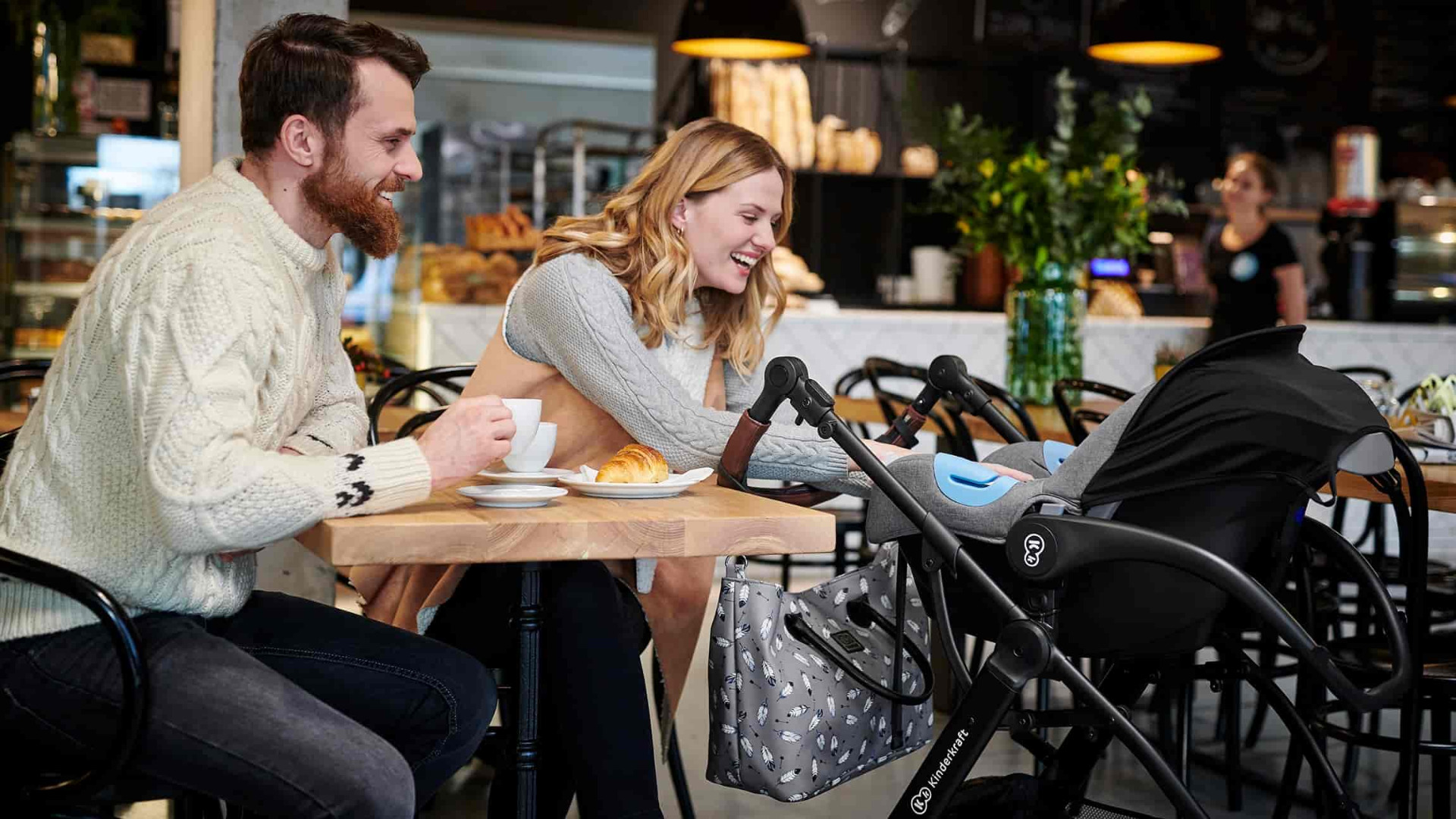 Śmiejąca się para siedząca w kawiarni. Kobieta sięga do wózka XMOOV. Mężczyzna trzyma w ręku filiżankę z kawą.