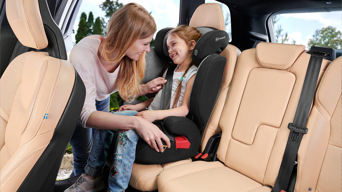Dziewczynka siedzi w samochodzie w foteliku samochodowym XPAND, mama zapina pasy bezpieczeństwa.