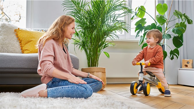 Mama śmieje się do dziecka, które jedzie na trójkołowym rowerku cutie kinderkraft. Są w domu, w tle widać zielone rośliny i szarą kanapę. Dziecko się uśmiecha.