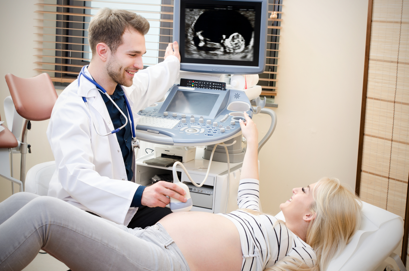 Lekarz w kitlu wykonuje badanie USG kobiecie ciężarnej. Pokazuje jej dziecko na monitorze, oboje są szczęśliwi.