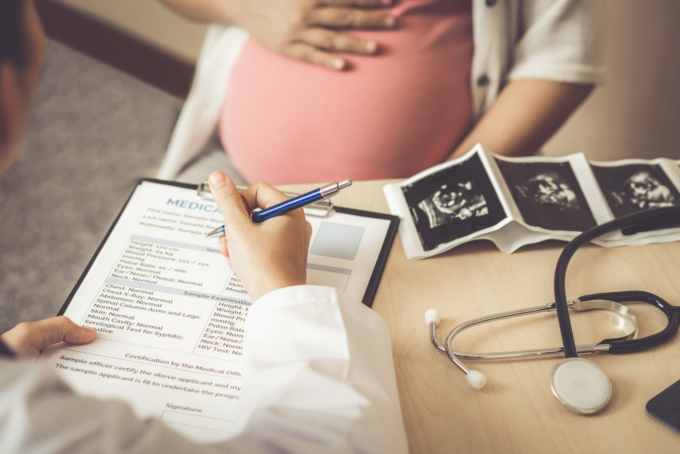 Lekarz ginekolog wypisuje skierowanie na badania w ciąży, w tle widać brzuszek ciążowy kobiety i wydruk z USG