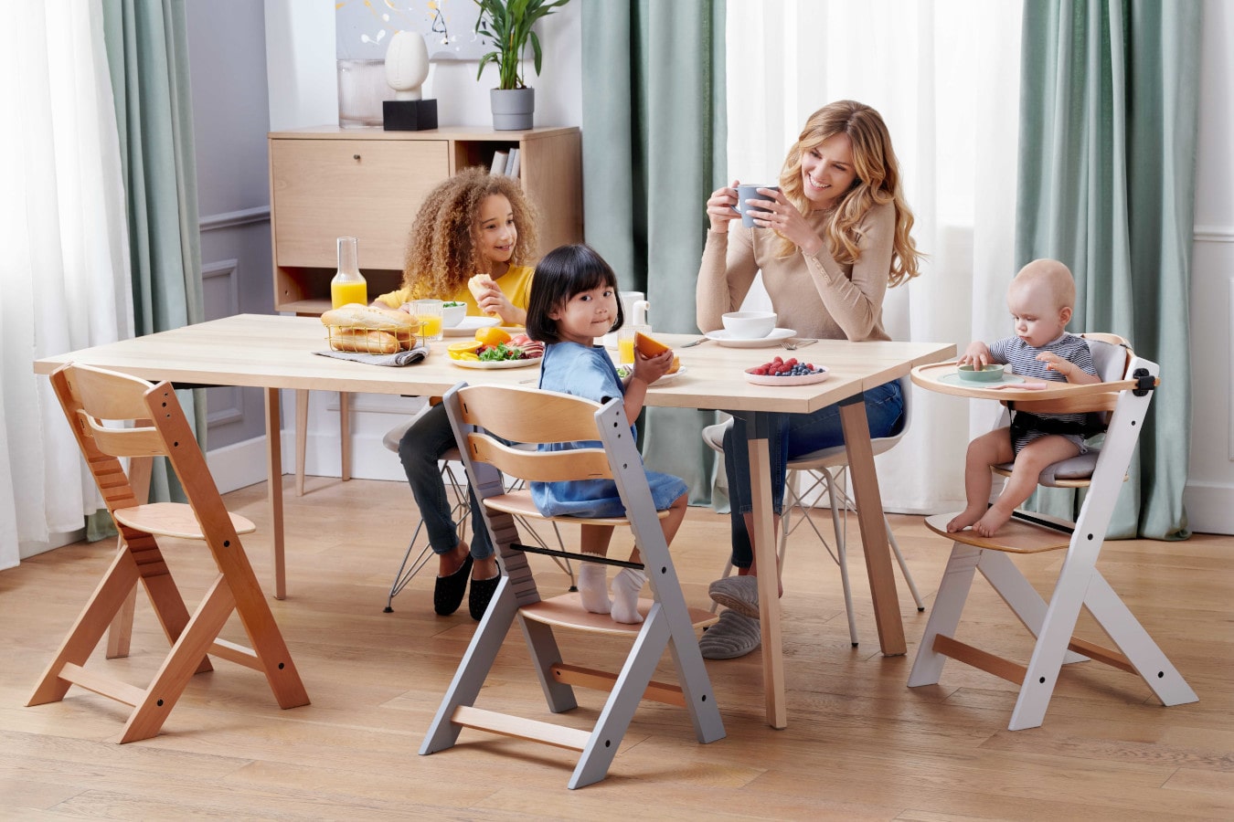 Mama i trójka dzieci siedzą przy jednym stole, mama pije kawę, dzieci są szczęśliwe. Krzesełko ENOCK jest w 3 konfiguracjach - od krzesła do karmienia po krzesło dla starszego dziecka.